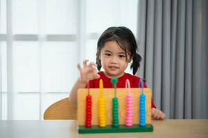 joven linda asiático bebé niña vistiendo rojo camiseta es aprendizaje el ábaco con de colores rosario a aprender cómo a contar en el mesa en el vivo habitación a hogar. niño bebé niña desarrollo estudiando concepto. foto