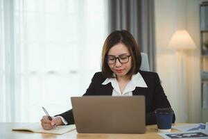 Asian businesswoman wearing suit and writing notes book and working laptop on table at home. Entrepreneur woman working for business at living room home. Business work home concept. photo
