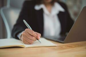 Close up of hand Asian businesswoman wearing suit and writing notes book and working laptop on table at home. Entrepreneur woman working for business at living room home. Business work home concept photo