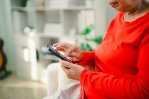 Happy senior old asian woman enjoying using mobile apps texting typing messages sit on sofa, smiling old lady holding smartphone looking smartphone browsing social media or learning technology at home photo