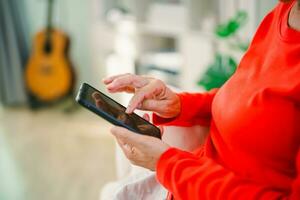 Happy senior old asian woman enjoying using mobile apps texting typing messages sit on sofa, smiling old lady holding smartphone looking smartphone browsing social media or learning technology at home photo