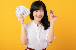 Portrait beautiful young asian woman enterpriser happy smile wearing white shirt and red plants holding cash dollar money and crypto digital currency isolation on yellow background. Wealth concept. photo