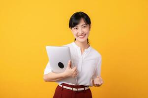 Portrait beautiful young asian woman enterpriser happy smile wearing white shirt and red plants showing confident body language gesture and holding laptop isolation on yellow background. photo