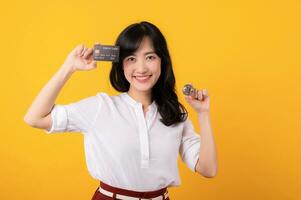 retrato hermosa joven asiático mujer emprendedor contento sonrisa vistiendo blanco camisa y rojo plantas participación crédito tarjeta y cripto digital moneda aislamiento en amarillo antecedentes. riqueza concepto. foto