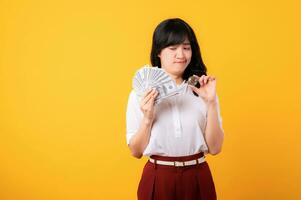 Portrait beautiful young asian woman enterpriser with doubt face wearing white shirt and red plants holding cash dollar money and crypto digital currency isolation on yellow background. Wealth concept photo