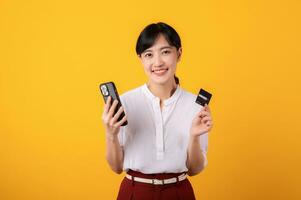 Portrait beautiful young asian woman enterpriser happy smile wearing white shirt and red plants holding credit card and smartphone isolation on yellow background. Online shopping concept. photo