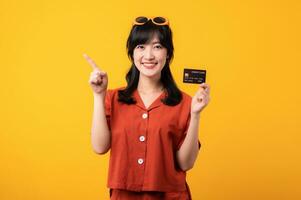 Portrait young beautiful asian woman happy smile dressed in orange clothes showing credit card and point finger to free space isolated on yellow background. Pay and purchase shopping payment concept. photo