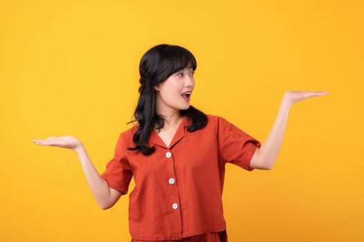 Portrait young beautiful asian woman happy smile dressed in orange clothes showing hand gesture, arm compare two variants demonstrate empty space isolated on yellow studio background. photo