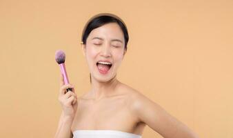 Portrait happy young asian woman with natural make up face holding cosmetic skin powder blusher isolated on beige background. Female apply skincare brush treatment. beauty product cosmetology concept. photo