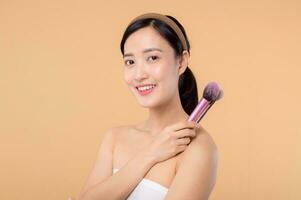 Portrait happy young asian woman with natural make up face holding cosmetic skin powder blusher isolated on beige background. Female apply skincare brush treatment. beauty product cosmetology concept. photo