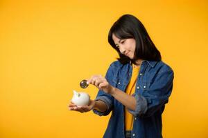 Unhappy asian young woman wearing yellow t-shirt denim shirt pulling digital coin crypto currency out of piggy bank isolated on yellow background. Payment digital money debt financial concept. photo