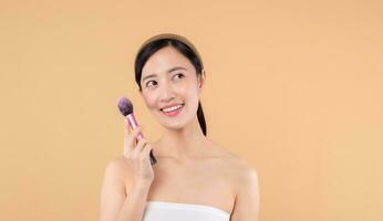 Portrait happy young asian woman with natural make up face holding cosmetic skin powder blusher isolated on beige background. Female apply skincare brush treatment. beauty product cosmetology concept. photo