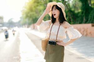 retrato de asiático joven mujer viajero con Costura sombrero y cesta y un cámara en verde público parque naturaleza antecedentes. viaje viaje estilo de vida, mundo viaje explorador o Asia verano turismo concepto. foto