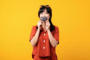 Portrait young beautiful asian woman happy smile dressed in orange clothes showing credit card isolated on yellow background. Pay and purchase shopping payment concept. photo