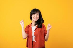 Portrait young beautiful asian woman happy smile dressed in orange clothes showing fist up hand gesture isolated on yellow studio background. Successful hooray celebrate young person concept. photo