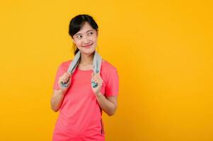 Portrait young asian sports fitness woman happy smile wearing pink sportswear and face towel doing exercise training workout against yellow studio background. wellbeing and healthy lifestyle concept. photo