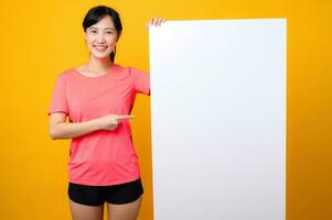 young asian sports fitness woman happy smile wearing pink sportswear standing behind the white blank banner or empty space advertisement board against yellow background. wellbeing lifestyle concept. photo