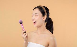 Portrait happy young asian woman with natural make up face holding cosmetic skin powder blusher isolated on beige background. Female apply skincare brush treatment. beauty product cosmetology concept. photo