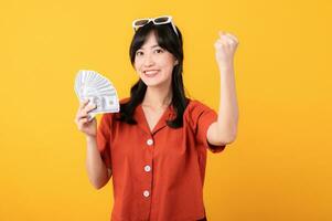 Portrait young beautiful asian woman happy smile dressed in orange clothes holding cash money and celebrate her success isolated on yellow background. Be rich concept. photo