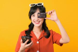 Portrait young beautiful asian woman happy smile dressed in orange clothes using smartphone and showing credit card isolated on yellow background. Pay and purchase shopping payment concept. photo