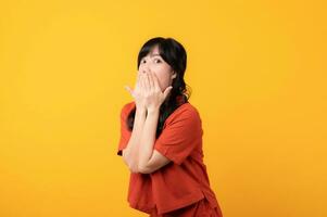 Portrait young beautiful asian woman happy smile dressed in orange clothes looking surprised, reacting amazed, raising eyebrows impressed isolated on yellow studio background. photo