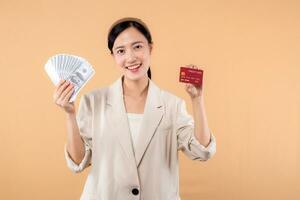 portrait of happy successful confident young asian business woman wearing white jacket holding cash money dollars and credit card standing over beige background. millionaire business, shopping concept photo