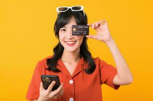 Portrait young beautiful asian woman happy smile dressed in orange clothes using smartphone and showing credit card isolated on yellow background. Pay and purchase shopping payment concept. photo