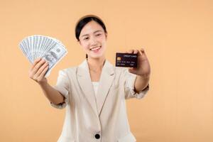 portrait of happy successful confident young asian business woman wearing white jacket holding cash money dollars and credit card standing over beige background. millionaire business, shopping concept photo