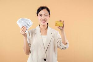 portrait of happy successful confident young asian business woman wearing white jacket holding cash money dollars and credit card standing over beige background. millionaire business, shopping concept photo