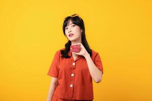 Portrait young beautiful asian woman happy smile dressed in orange clothes showing credit card isolated on yellow background. Pay and purchase shopping payment concept. photo