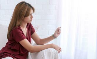 Worried young woman holding checking pregnancy test result sitting on white sofa in living room at home. photo
