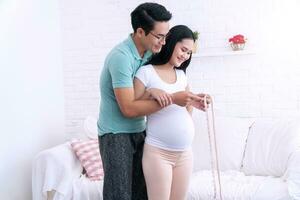 Happy young man measuring the waist of the pregnant his wife standing in living room. Husband measuring waist circumference his wife belly at home. photo