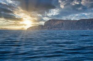 ver desde el mar a el Oeste capa en Noruega en Brillo Solar y pesado nubes paisaje foto