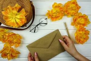 Women's hands are holding a craft envelope. On a white wooden table there is a wicker bag and yellow terry tulips. photo