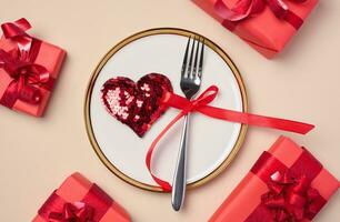 Red heart on a round white plate and fork on a beige background, top view. Gifts all around photo