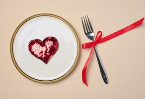 Red heart on a round white plate and fork on a beige background, top view photo