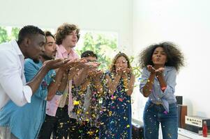 Multi-ethnic group of friends celebrating party and having fun blowing coloured confetti at home. photo