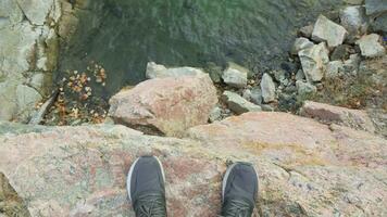 Legs of a man standing on the edge of a precipice and looking down at the rocky shore. video