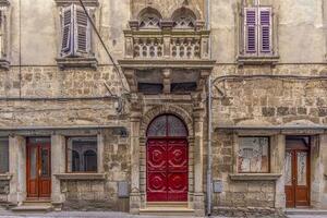 Image of a red entrance door to a residential building with a stone balcony and window with shutters on a antique facade photo