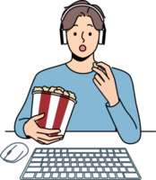 Man with popcorn watches movie sitting at table with computer and looks at screen intrigued png