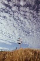 Cross on top of a hill against a blue sky with white clouds. Silhouette of a cross in the field at sunset with dramatic sky. photo