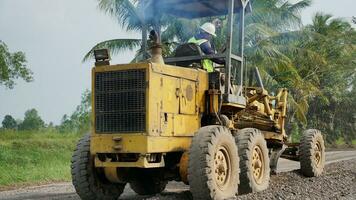 a yellow motor grader leveling stones and gravel for road repairs in Lampung. Behind view. photo