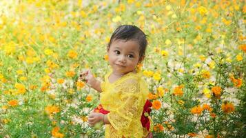 pequeño linda niña vistiendo amarillo balinés vestir jugando en amarillo y blanco flor jardín foto