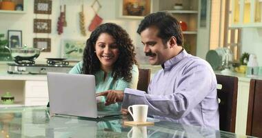 Indian couple sitting at dining table at home in kitchen having fun while shopping online video