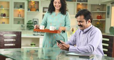 Video of Couple In Kitchen, Wife Bringing Coffee While Husband Typing on mobile sitting on Kitchen Table