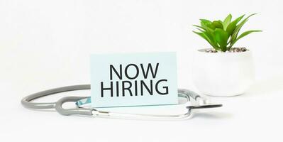 NOW HIRING word on notebook, stethoscope and green plant photo
