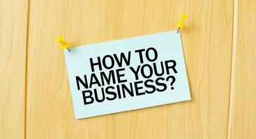HOW TO NAME YOUR BUISINESS sign written on sticky note pinned on wooden wall photo