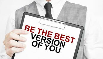 BE THE BEST VERSION OF YOU inscription on a notebook in the hands of a businessman on a gray background, a man points with a finger to the text photo