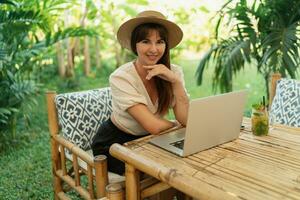 Pretty woman in straw hat using lap top while traveling in asia.  Working remotely in stylish tropical cafe. photo