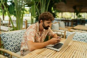 Handsome man with beard using lap top and mobyle phone , sitting in outdoor  cafe with tropical interior. photo